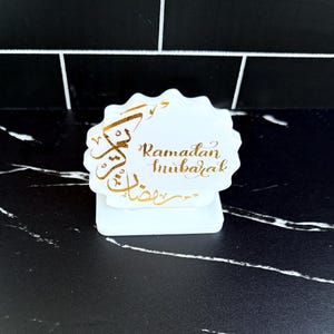 May include: A white, scalloped-edge table decoration with gold calligraphy that reads "Ramadan Mubarak." The decoration sits on a white base, and is set against a black and white tiled background.