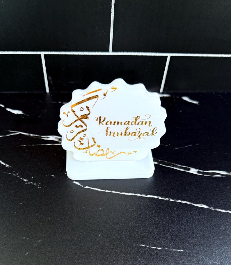 May include: A white, scalloped-edge table decoration with gold calligraphy that reads "Ramadan Mubarak." The decoration sits on a white base, and is set against a black and white tiled background.