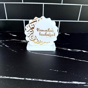 May include: A white, scalloped-edge Ramadan Mubarak decoration with gold calligraphy. The decoration is on a small rectangular base. The background is a black and white tiled wall and a black marble surface.