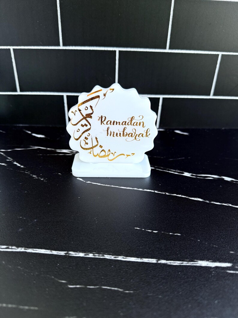 May include: A white, scalloped-edge Ramadan Mubarak decoration with gold calligraphy. The decoration is on a small rectangular base. The background is a black and white tiled wall and a black marble surface.