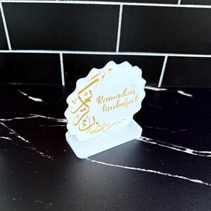 May include: A white, scalloped-edge Ramadan Mubarak decoration with gold calligraphy. The decoration is on a white base and sits on a black and white surface. The text is in gold and reads "Ramadan Mubarak".