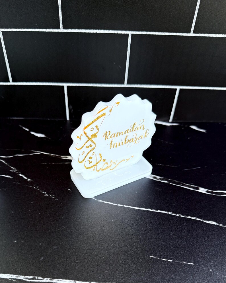 May include: A white, scalloped-edge Ramadan Mubarak decoration with gold calligraphy. The decoration is on a white base and sits on a black and white surface. The text is in gold and reads "Ramadan Mubarak".