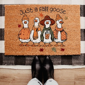May include: A rectangular tan coir doormat featuring a graphic illustration of four geese wearing autumn-themed clothing and accessories. The text "Just a fall goose" is printed in black above the geese, surrounded by scattered orange and red leaves.