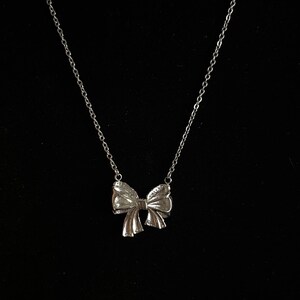 Puede incluir: Un collar plateado con un colgante de lazo. El lazo presenta detalles texturizados y cuelga de una delicada cadena. El collar se muestra sobre un fondo negro.