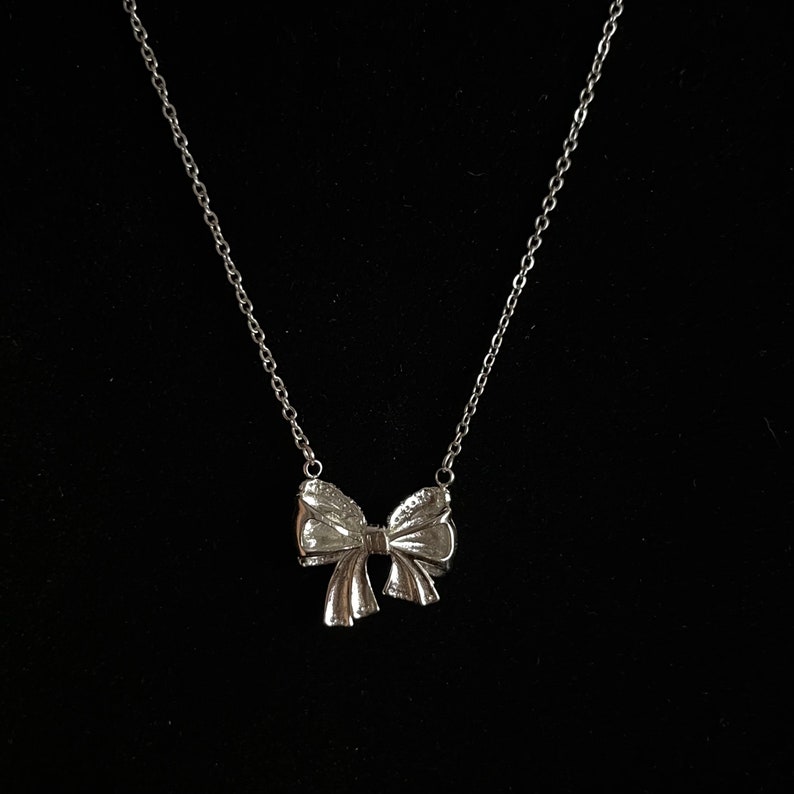 Puede incluir: Un collar plateado con un colgante de lazo. El lazo presenta detalles texturizados y cuelga de una delicada cadena. El collar se muestra sobre un fondo negro.