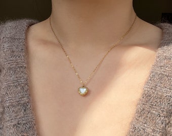 Puede incluir: Un collar de cadena dorada con un pequeño colgante de corazón blanco. El corazón tiene un diseño detallado y adornado alrededor del borde.