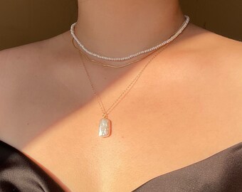 Puede incluir: Un collar de varios niveles con una cadena de oro y un colgante de perla blanca grande. La capa superior es una cadena delicada de pequeñas perlas blancas.