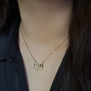 Puede incluir: Un collar dorado con un colgante de lazo. El lazo tiene un dise&ntilde;o texturizado y est&aacute; adornado con peque&ntilde;os detalles brillantes. La cadena del collar es delicada y dorada. El fondo es de un color oscuro y s&oacute;lido.