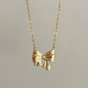Puede incluir: Un collar dorado con un colgante de lazo. El lazo tiene una textura detallada y cuelga de una delicada cadena. El collar se muestra sobre un fondo neutro, resaltando el dise&ntilde;o de la joya.