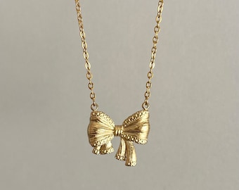 May include: A gold-colored necklace featuring a bow pendant. The bow has detailed texturing and is suspended from a delicate chain. The necklace is displayed against a neutral background, highlighting the jewelry's design.