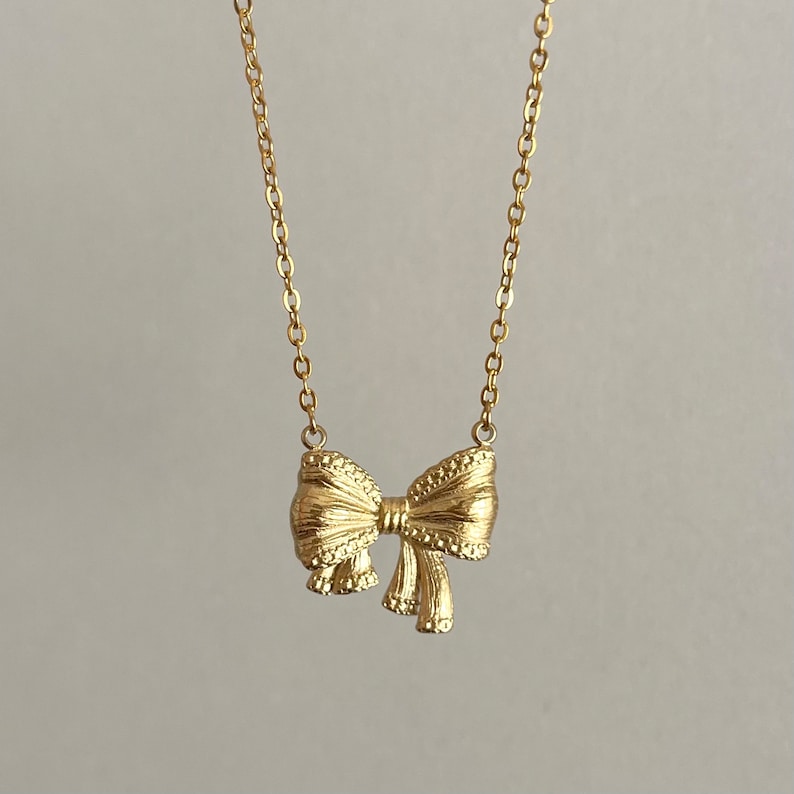Puede incluir: Un collar dorado con un colgante de lazo. El lazo tiene una textura detallada y cuelga de una delicada cadena. El collar se muestra sobre un fondo neutro, resaltando el dise&ntilde;o de la joya.