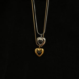Può includere: Una collana d'argento e oro con due ciondoli a forma di cuore. Il ciondolo superiore è d'argento con un cuore nero all'interno, e il ciondolo inferiore è d'oro con un cuore nero all'interno.