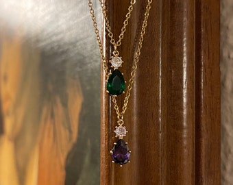 Puede incluir: Un collar de cadena de oro con dos colgantes en forma de lágrima. El colgante superior es verde con un acento de piedra clara. El colgante inferior es morado con un acento de piedra clara.