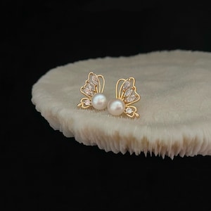 Könnte beinhalten: Ein Paar goldene Schmetterlings-Ohrringe mit weißen Perlenakzenten. Die Ohrringe befinden sich auf einem weißen Hintergrund.