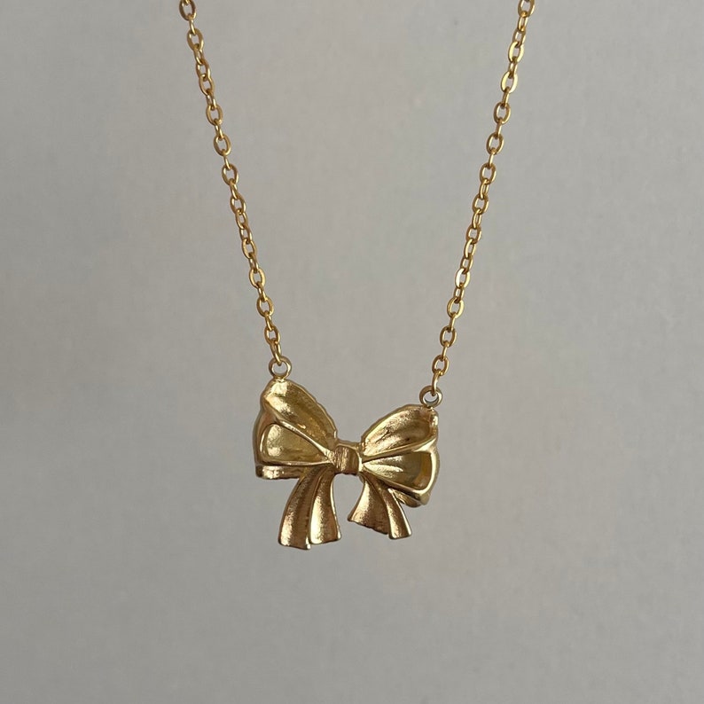Puede incluir: Un collar dorado con un colgante de lazo. El lazo presenta pliegues texturizados y cuelga de una delicada cadena. El collar se muestra sobre un fondo neutro, resaltando el dise&ntilde;o de la joya.