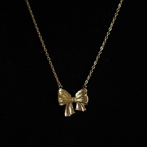 Puede incluir: Un collar dorado con un colgante de lazo. El lazo tiene superficies texturizadas y cuelga de una delicada cadena. El collar se muestra sobre un fondo negro.