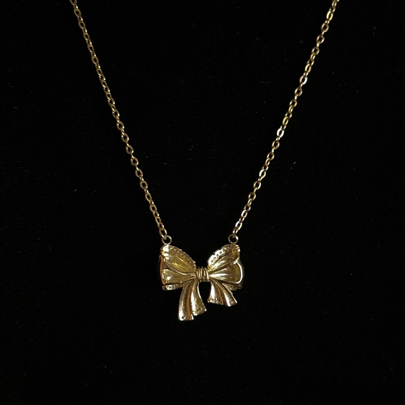 Puede incluir: Un collar dorado con un colgante de lazo. El lazo tiene superficies texturizadas y cuelga de una delicada cadena. El collar se muestra sobre un fondo negro.