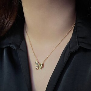 Puede incluir: Un collar dorado con un colgante de lazo. El lazo tiene detalles intrincados y cuelga de una delicada cadena. El collar se muestra sobre un fondo oscuro, resaltando su brillo y dise&ntilde;o.