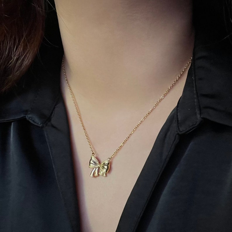 Puede incluir: Un collar dorado con un colgante de lazo. El lazo tiene detalles intrincados y cuelga de una delicada cadena. El collar se muestra sobre un fondo oscuro, resaltando su brillo y dise&ntilde;o.