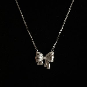 Puede incluir: Un collar plateado con un colgante de lazo. El lazo tiene detalles intrincados y cuelga de una cadena fina. El collar se muestra sobre un fondo negro.