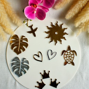 Könnte beinhalten: Ein Satz goldener und silberner Acrylformen, darunter eine Sonne, eine Schildkröte, ein Herz, eine Krone, ein Blatt und ein Flugzeug. Die Formen befinden sich auf einem weißen Hintergrund.