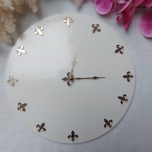 Op de afbeelding: Witte ronde klok met gouden fleur-de-lis accenten rond de rand en gouden wijzers.