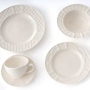 May include: A cream-coloured ceramic dinnerware set comprising a large plate, a bowl, a smaller plate, a teacup and a saucer. The rims of the plates and bowl have textured patterns, and the teacup and saucer have a matching design.