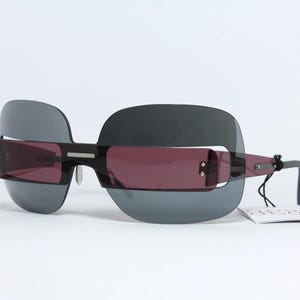 May include: Unique sunglasses with a black frame and dark gray lenses. The sunglasses have a rectangular shape with a dark red tint on the inner lenses. A white tag is attached to the frame.