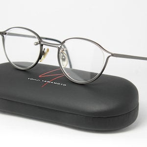 May include: Silver-toned metal eyeglasses with round lenses and a black case with the text "Yohji Yamamoto" printed on it.