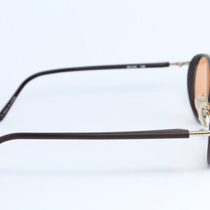 May include: Brown-framed sunglasses with a gold-colored bridge and temples. The lenses are a light orange color. The arms are a dark brown color. The glasses are set against a white background.