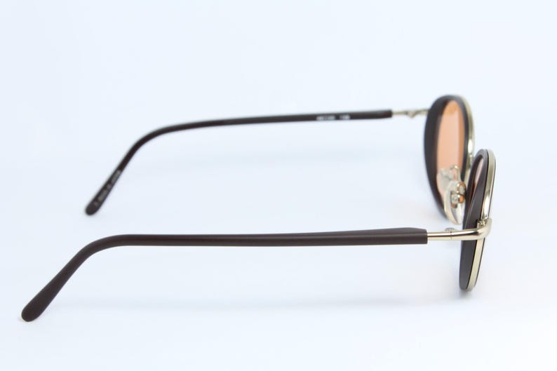 May include: Brown-framed sunglasses with a gold-colored bridge and temples. The lenses are a light orange color. The arms are a dark brown color. The glasses are set against a white background.