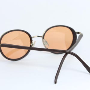 May include: Round sunglasses with brown frames and orange-tinted lenses. The metal bridge and hinges provide a contrast to the dark brown arms. The sunglasses are set against a white background.