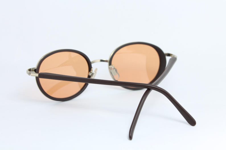 May include: Round sunglasses with brown frames and orange-tinted lenses. The metal bridge and hinges provide a contrast to the dark brown arms. The sunglasses are set against a white background.