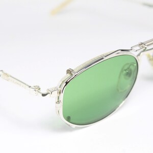 May include: Silver-framed sunglasses with green lenses. The glasses have a double bridge and a classic aviator style. The temples are thin and silver, and the lenses are a vibrant green color. The overall design is sleek and modern.