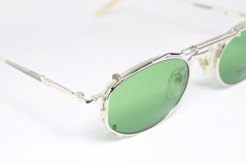 May include: Silver-framed sunglasses with green lenses. The glasses have a double bridge and a classic aviator style. The temples are thin and silver, and the lenses are a vibrant green color. The overall design is sleek and modern.