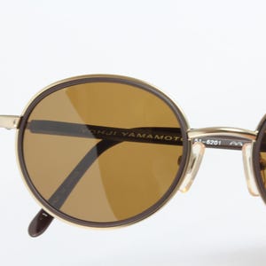 K&ouml;nnte beinhalten: Runde Sonnenbrille mit braun get&ouml;nten Gl&auml;sern und einem goldfarbenen Rahmen. Die Sonnenbrille hat einen dunkelbraunen Rand um die Gl&auml;ser und der Text "YOHJI YAMAMOTO" ist auf dem Rahmen sichtbar. Die B&uuml;gel sind dunkelbraun.