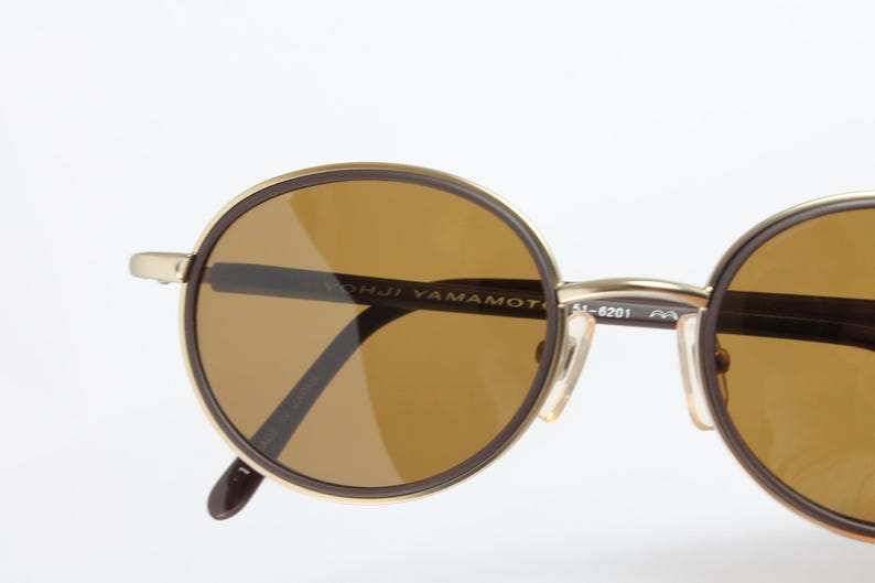 K&ouml;nnte beinhalten: Runde Sonnenbrille mit braun get&ouml;nten Gl&auml;sern und einem goldfarbenen Rahmen. Die Sonnenbrille hat einen dunkelbraunen Rand um die Gl&auml;ser und der Text "YOHJI YAMAMOTO" ist auf dem Rahmen sichtbar. Die B&uuml;gel sind dunkelbraun.
