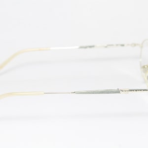 May include: Silver-toned eyeglasses with a classic oval frame design. The temples and frame are silver, with light beige earpieces. The glasses are shown in a side profile view against a white background.