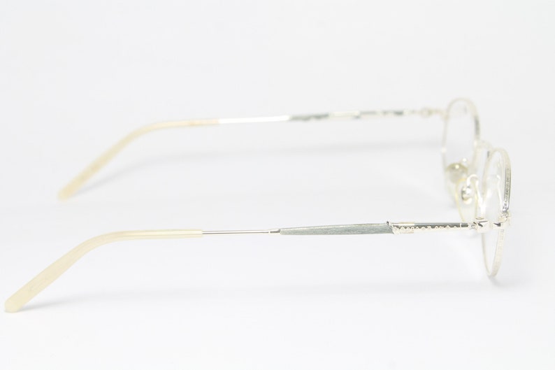 May include: Silver-toned eyeglasses with a classic oval frame design. The temples and frame are silver, with light beige earpieces. The glasses are shown in a side profile view against a white background.