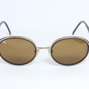 K&ouml;nnte beinhalten: Ovale Sonnenbrille mit braunen Gl&auml;sern und einem goldfarbenen Rahmen. Die B&uuml;gel sind dunkelbraun. Die Sonnenbrille ist vor einem wei&szlig;en Hintergrund platziert.