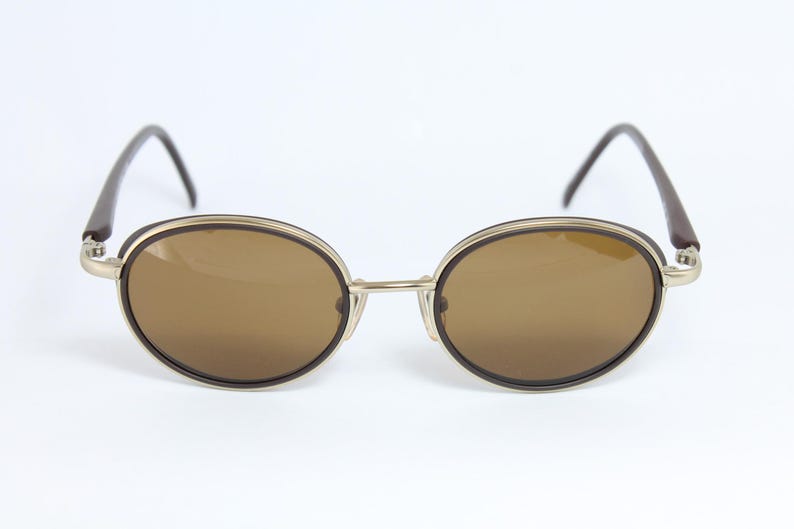 K&ouml;nnte beinhalten: Ovale Sonnenbrille mit braunen Gl&auml;sern und einem goldfarbenen Rahmen. Die B&uuml;gel sind dunkelbraun. Die Sonnenbrille ist vor einem wei&szlig;en Hintergrund platziert.