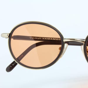 May include: Close-up of round sunglasses with orange-tinted lenses. The frames are gold-colored with a black outline. The brand name "YOHJI YAMAMOTO" is printed on the frame. The sunglasses are made in Japan.