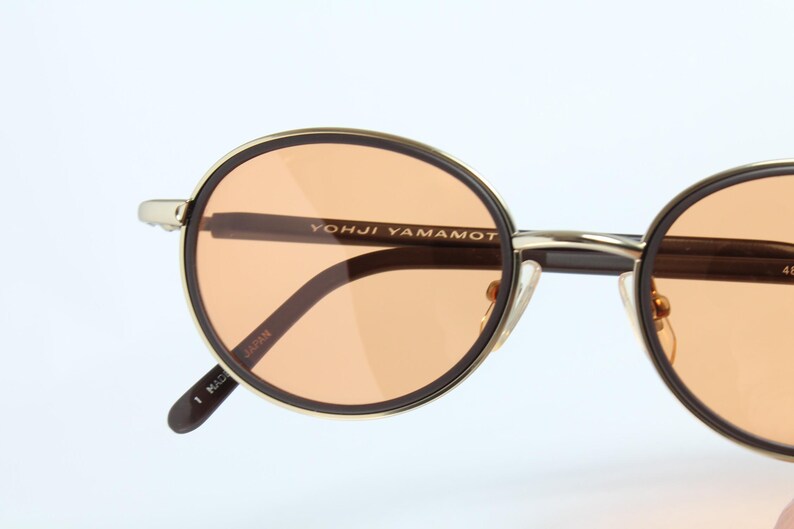 May include: Close-up of round sunglasses with orange-tinted lenses. The frames are gold-colored with a black outline. The brand name "YOHJI YAMAMOTO" is printed on the frame. The sunglasses are made in Japan.