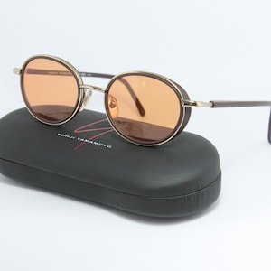 May include: Round sunglasses with brown frames and orange lenses rest on a black case. The sunglasses have a gold-colored bridge and arms. The case has the brand name "YOHJI YAMAMOTO" in red.