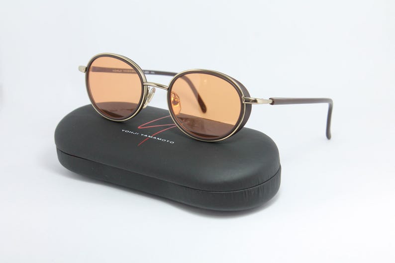 May include: Round sunglasses with brown frames and orange lenses rest on a black case. The sunglasses have a gold-colored bridge and arms. The case has the brand name "YOHJI YAMAMOTO" in red.