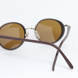 K&ouml;nnte beinhalten: Runde Sonnenbrille mit braunen Gl&auml;sern und dunkelbraunen B&uuml;geln. Die Fassungen sind hellmetallfarben. Die Sonnenbrille ist vor einem wei&szlig;en Hintergrund platziert.