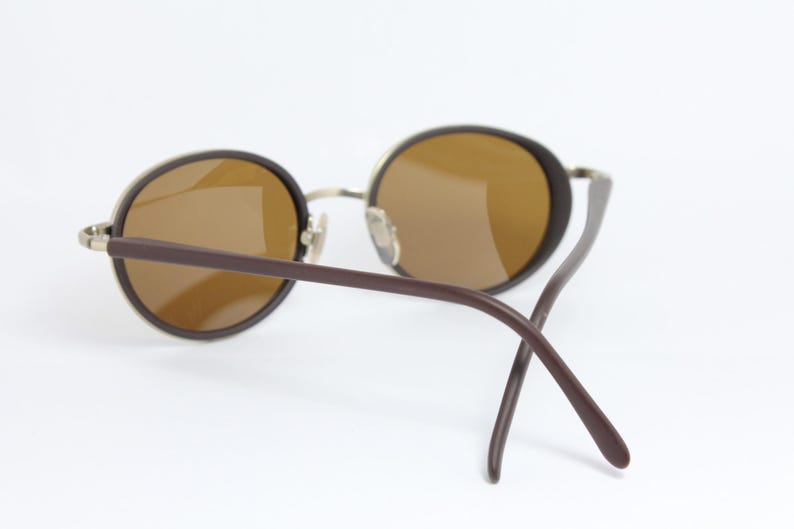 K&ouml;nnte beinhalten: Runde Sonnenbrille mit braunen Gl&auml;sern und dunkelbraunen B&uuml;geln. Die Fassungen sind hellmetallfarben. Die Sonnenbrille ist vor einem wei&szlig;en Hintergrund platziert.