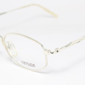 May include: Close-up of Matsuda eyeglasses with a silver metal frame. The glasses have a rounded rectangular shape with a thin frame and arms. The brand name "MATSUDA" is printed on the lens.