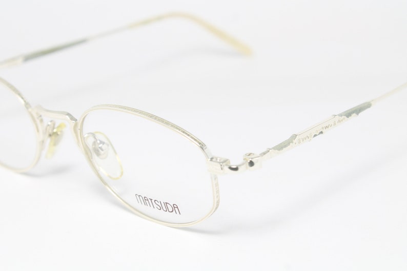 May include: Close-up of Matsuda eyeglasses with a silver metal frame. The glasses have a rounded rectangular shape with a thin frame and arms. The brand name "MATSUDA" is printed on the lens.