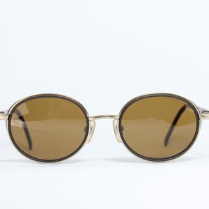 K&ouml;nnte beinhalten: Runde Sonnenbrille mit braunen Gl&auml;sern und einem goldfarbenen Metallrahmen. Die Gl&auml;ser sind von einem dunkelbraunen Rand umgeben. Die B&uuml;gel sind d&uuml;nn und goldfarben. Die Sonnenbrille ist vor einem wei&szlig;en Hintergrund platziert.